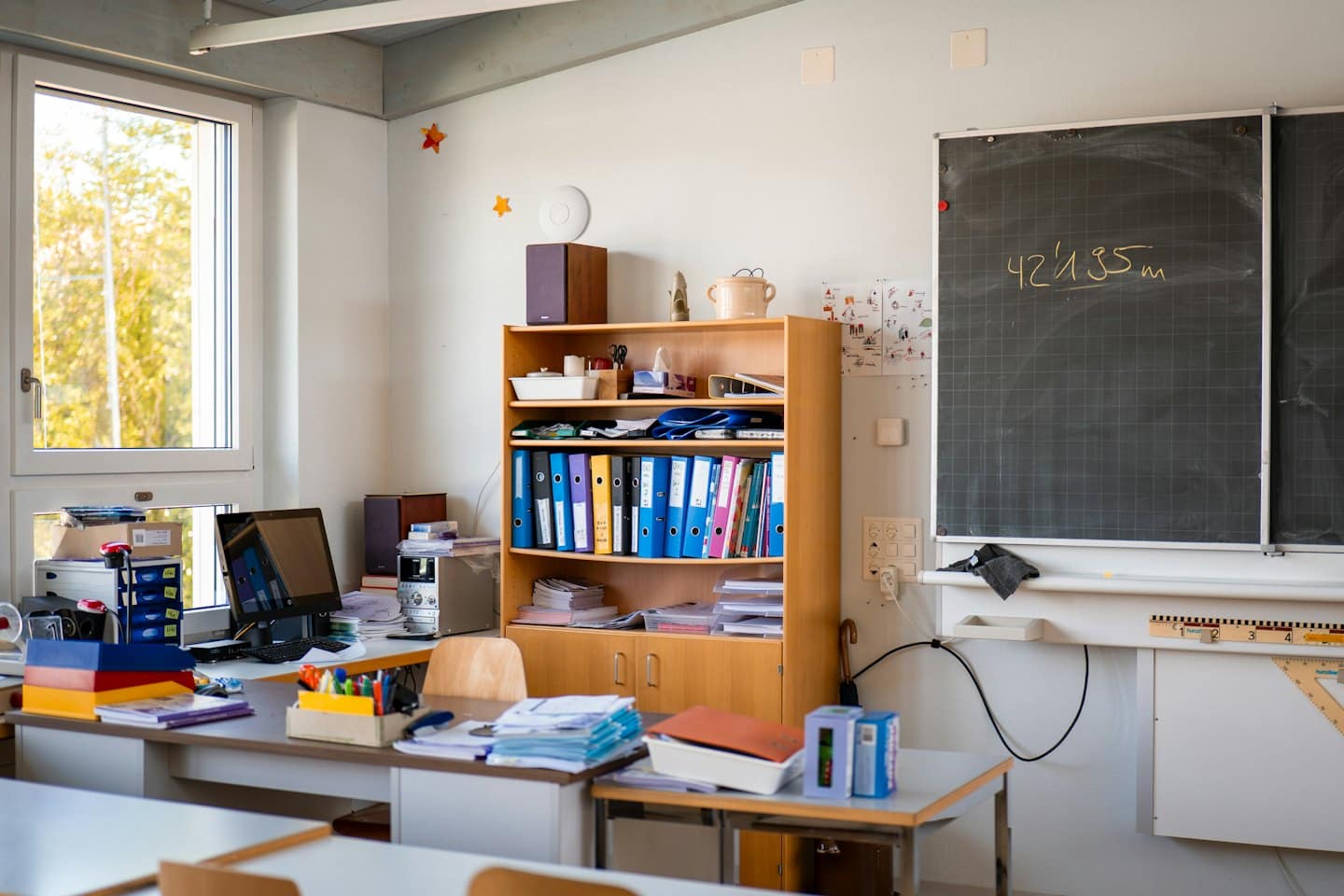 Classroom desk with computer and supplies near blackboard and drawings.